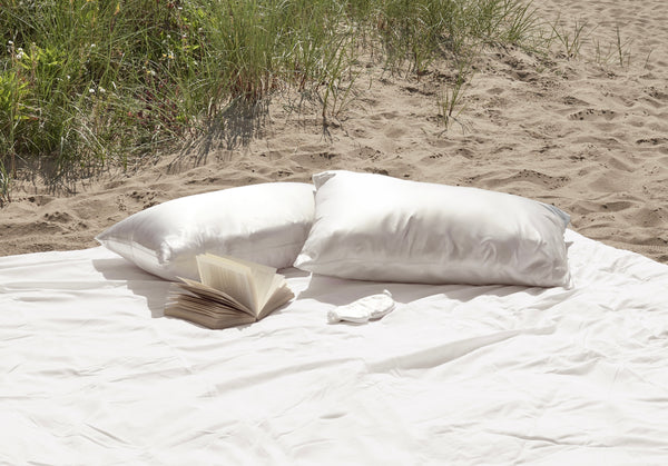 two ivory silk pillowcases laid down on a white duvet on the sand 