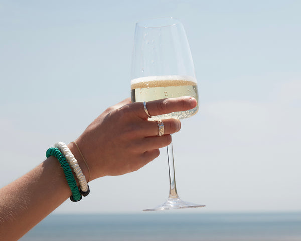 Lady holding a glass of champagne with 2 silk scrunchies on wrist, blue sea and sky background