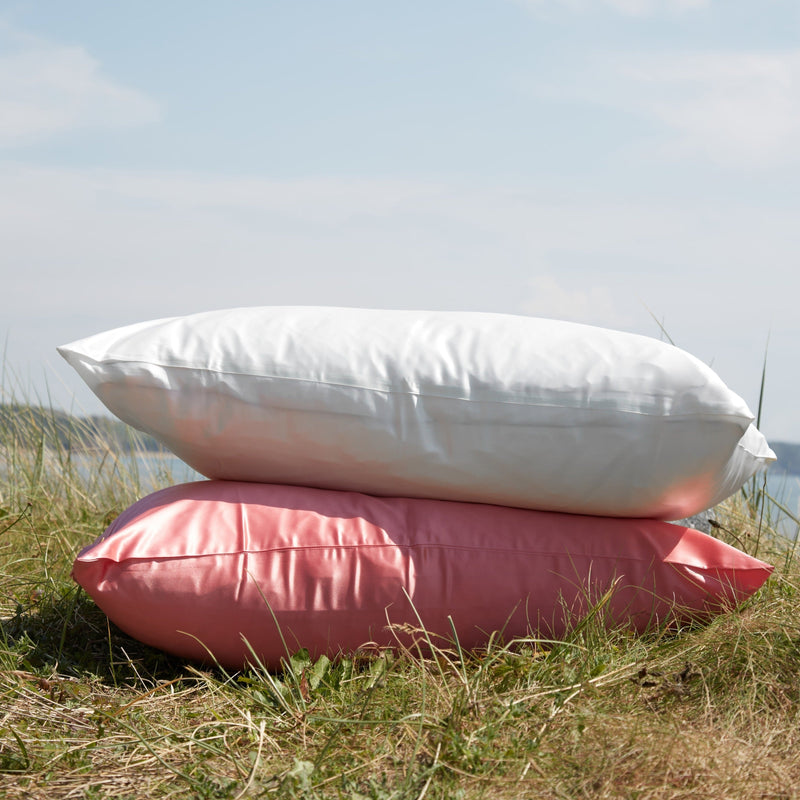 Two mulberry silk pillowcases on top of each other sitting on the grass. One pillowcase is ivory and the other is watermelon coloured.